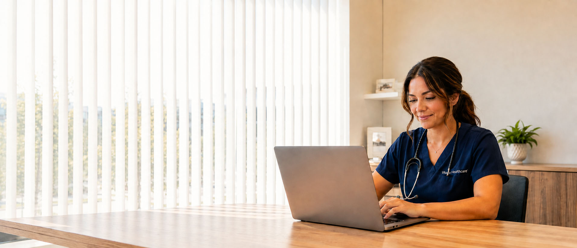 A nurse practitioner in navy scrubs works at a laptop in a bright clinic office.